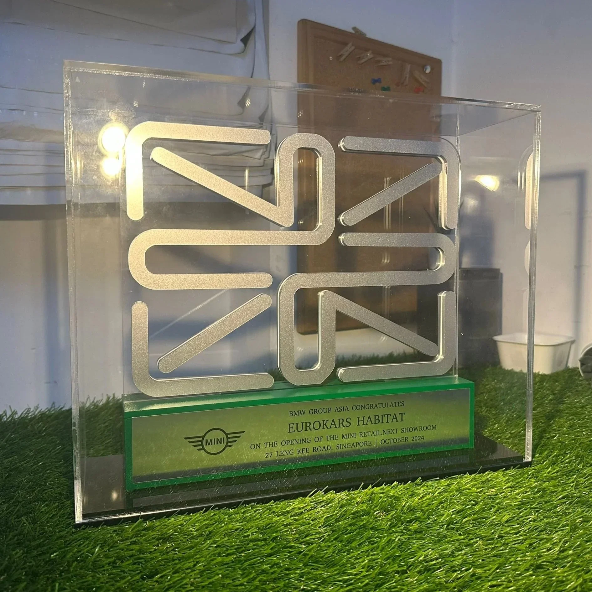 A transparent award trophy with a silver MINI logo and a green plaque at the bottom. The plaque has text congratulating Eurokars Habitat on the opening of a MINI showroom in Singapore, dated October 2024, and features a small MINI logo.