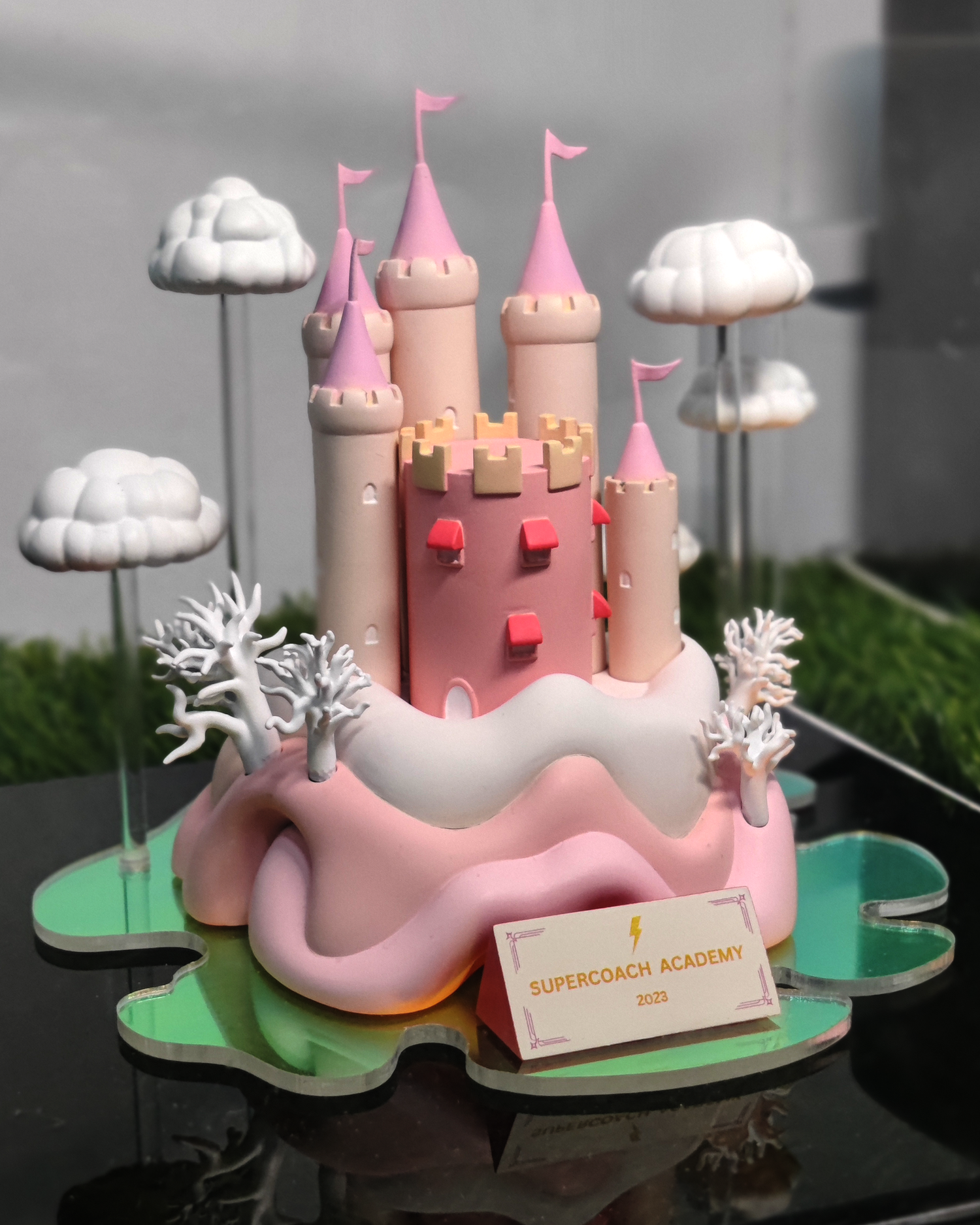 A pink castle model with multiple towers and flags, surrounded by white clouds and trees, on a green base with a sign reading 'Supercoach Academy 2023'.
