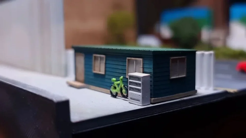 Miniature model of a blue house with a small green bicycle and gray storage chest outside on the sidewalk.