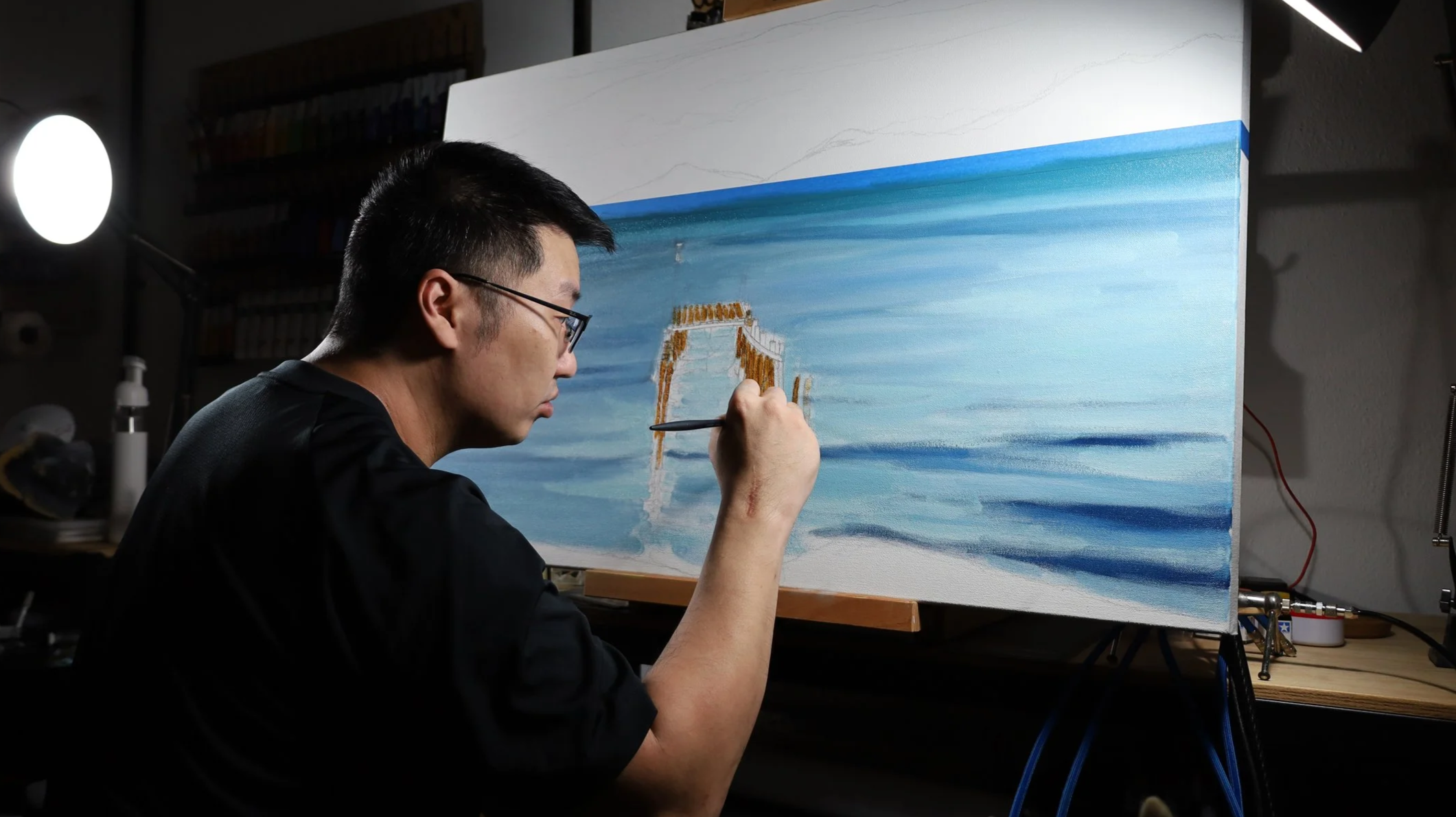 Artist painting a seascape with a bridge on a large canvas in a studio, surrounded by lighting and art supplies.