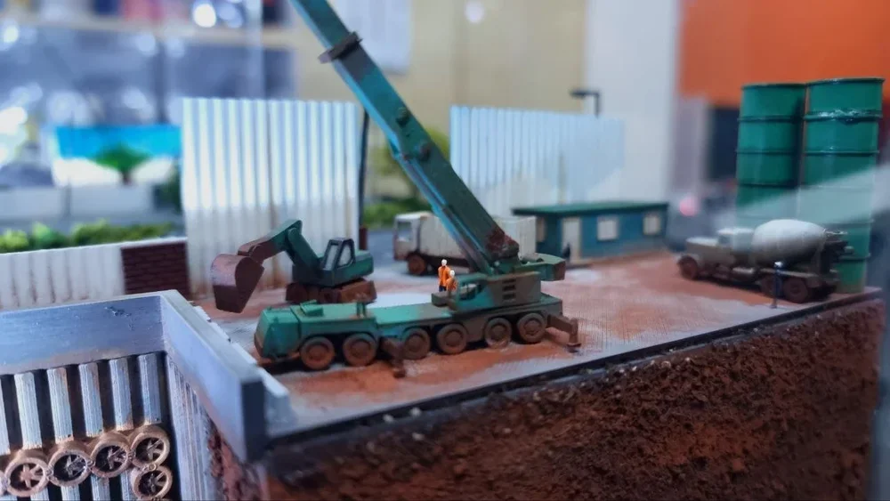 Miniature model of a construction site with a green crane, a small excavator, a cement mixer truck, and green storage containers.