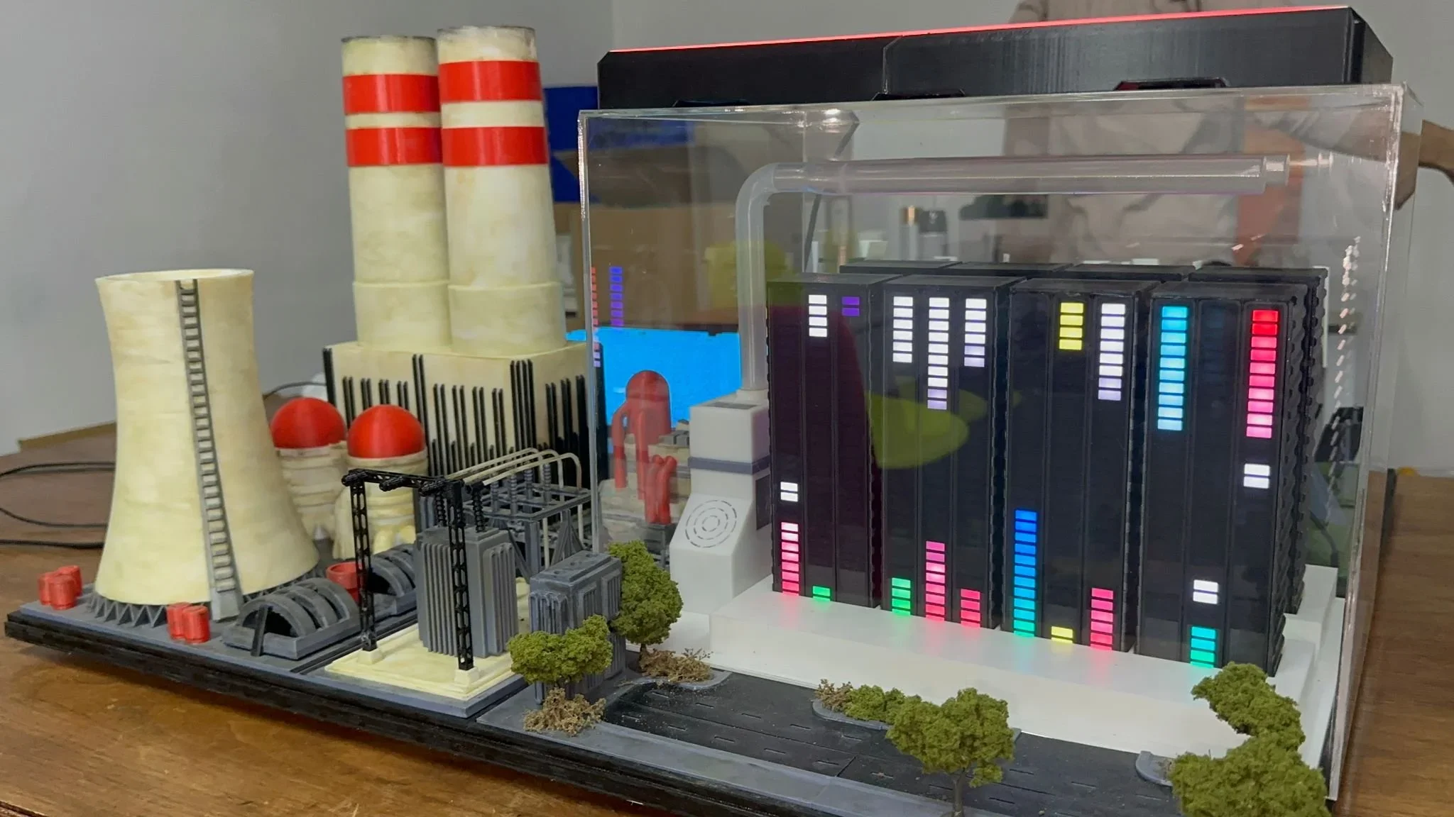 Miniature model of a nuclear power plant with cooling towers, cooling pools, and cooling rods, surrounded by small trees and infrastructure, some displays with illuminated colorful bars.