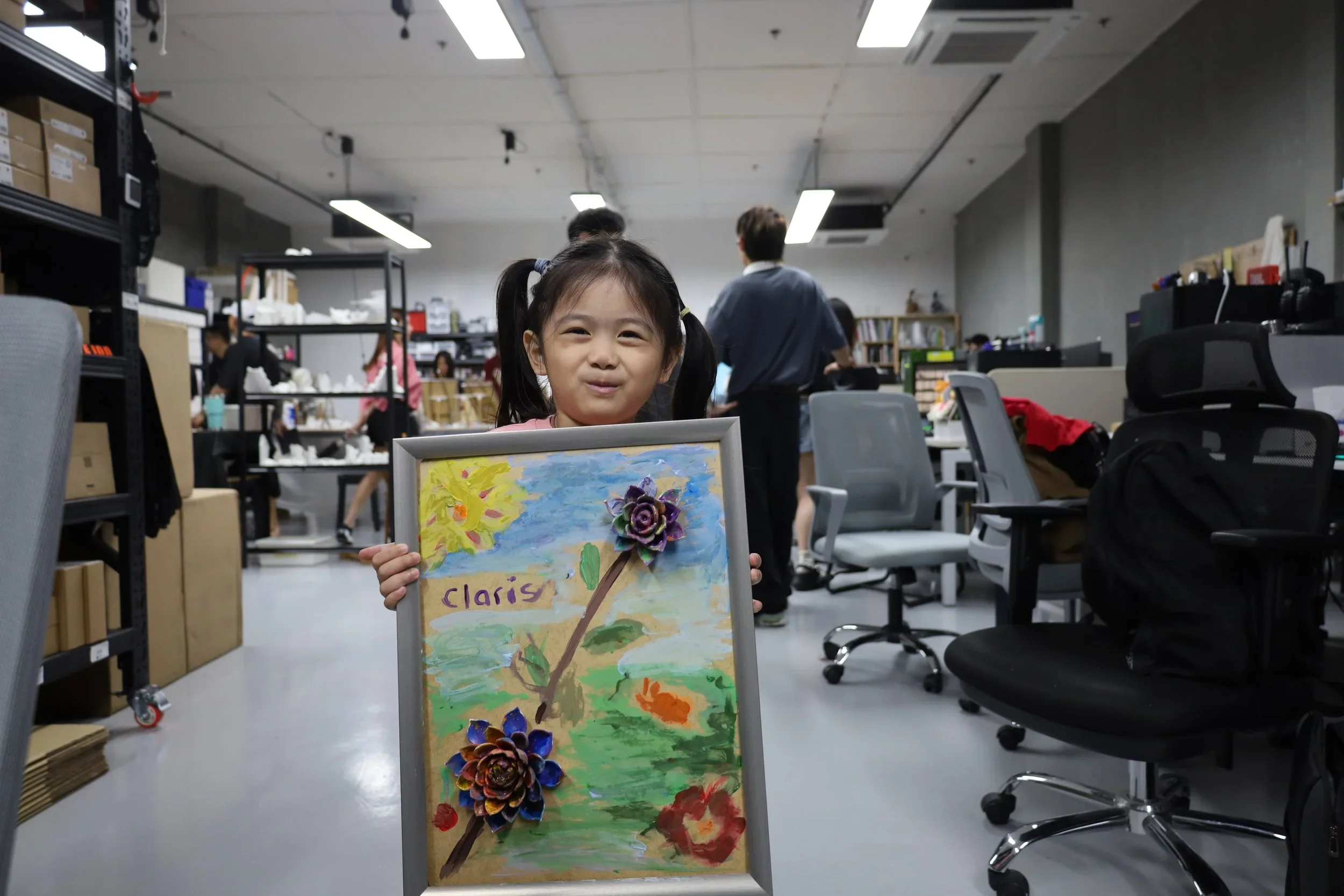 A young girl with pigtails holding a framed painting with the name 'Clarise' on it, featuring colorful flowers and a blue sky, inside a busy art classroom or studio with chairs, shelves, and other people working in the background.