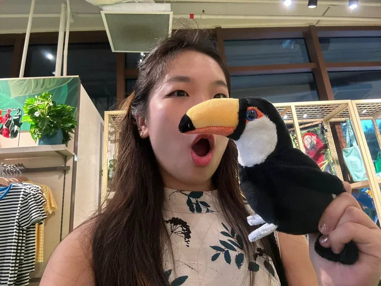 A woman is holding a plush toucan toy near her face, with her mouth open in surprise. She is inside a store with shelves and clothing in the background.