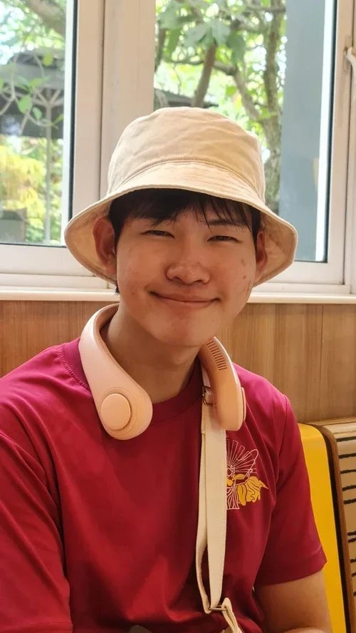 A young person wearing a beige bucket hat, pink headphones around their neck, and a maroon shirt, smiling indoors with a window and greenery in the background.