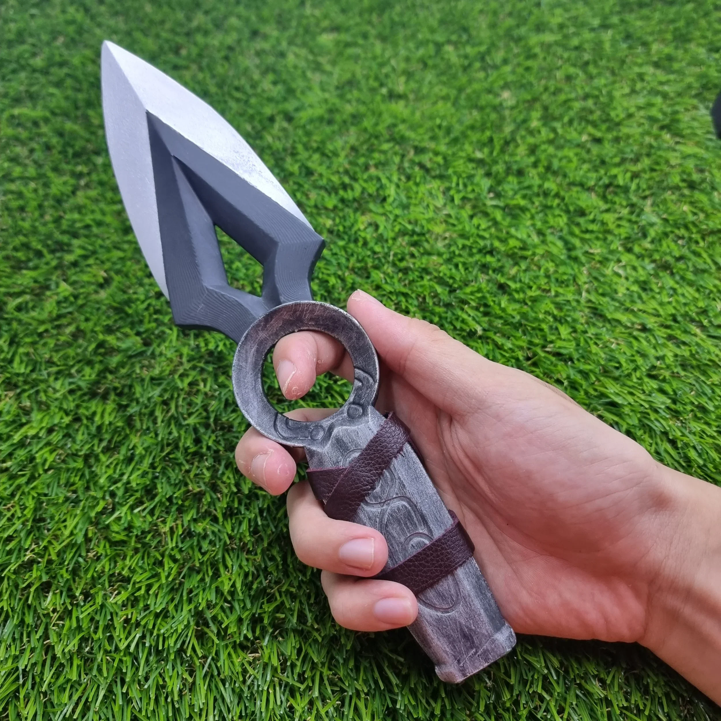A person holding a toy or replica sword with a black and gray blade and a gray handle with a leather strap, over a background of green artificial grass.