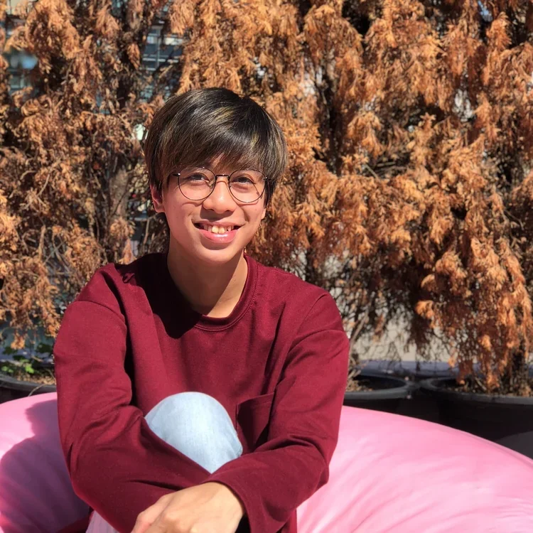 A young person with glasses and a maroon long-sleeve shirt, smiling with a gap-tooth, sitting in front of a bush with brown, dried leaves, on a pink inflatable lounger outside.