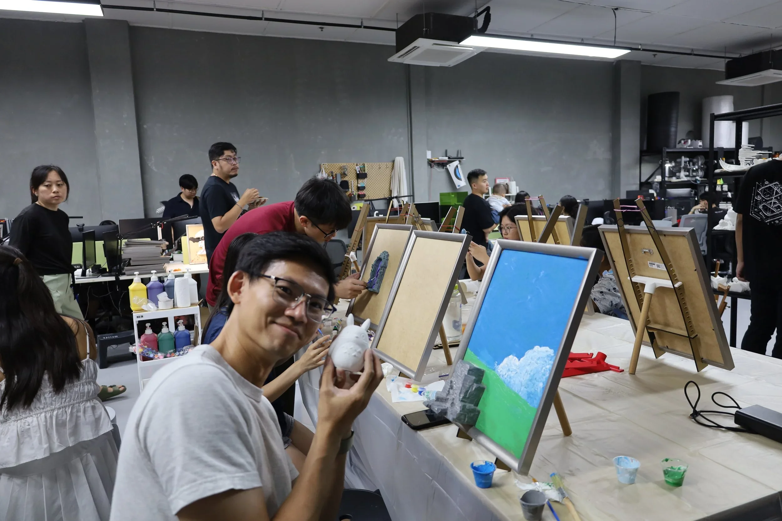 Group of people participating in an indoor painting class, with several easels, paints, and canvases, some completed artworks visible, and a man in the foreground holding a ceramic head model.