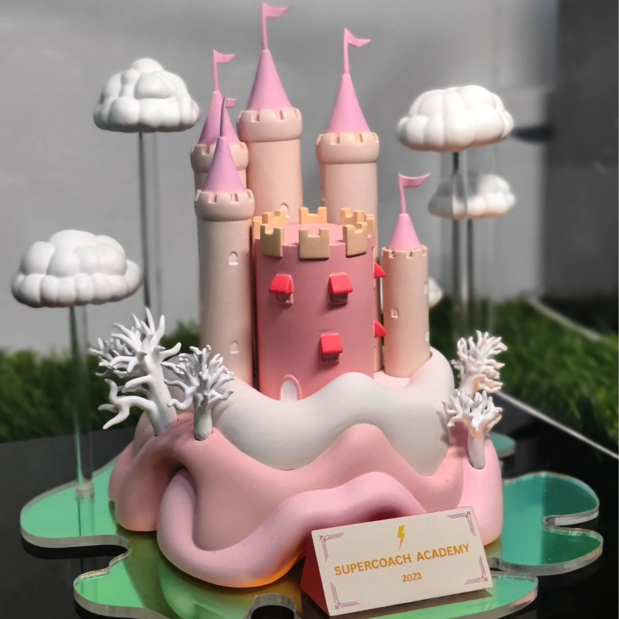 A decorative cake model resembling a pink castle with towers, flags, and trees, labeled 'Supercoach Academy 2023'.