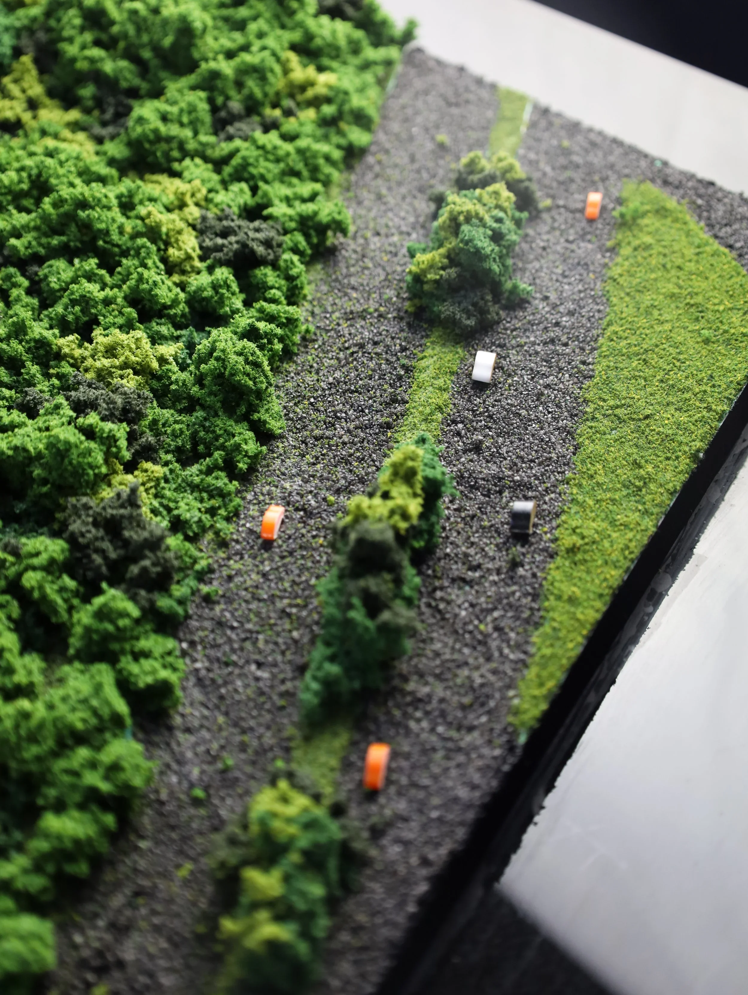Miniature model of a forested landscape with trees, grass, rocks, and small toy vehicles on a gravel road.