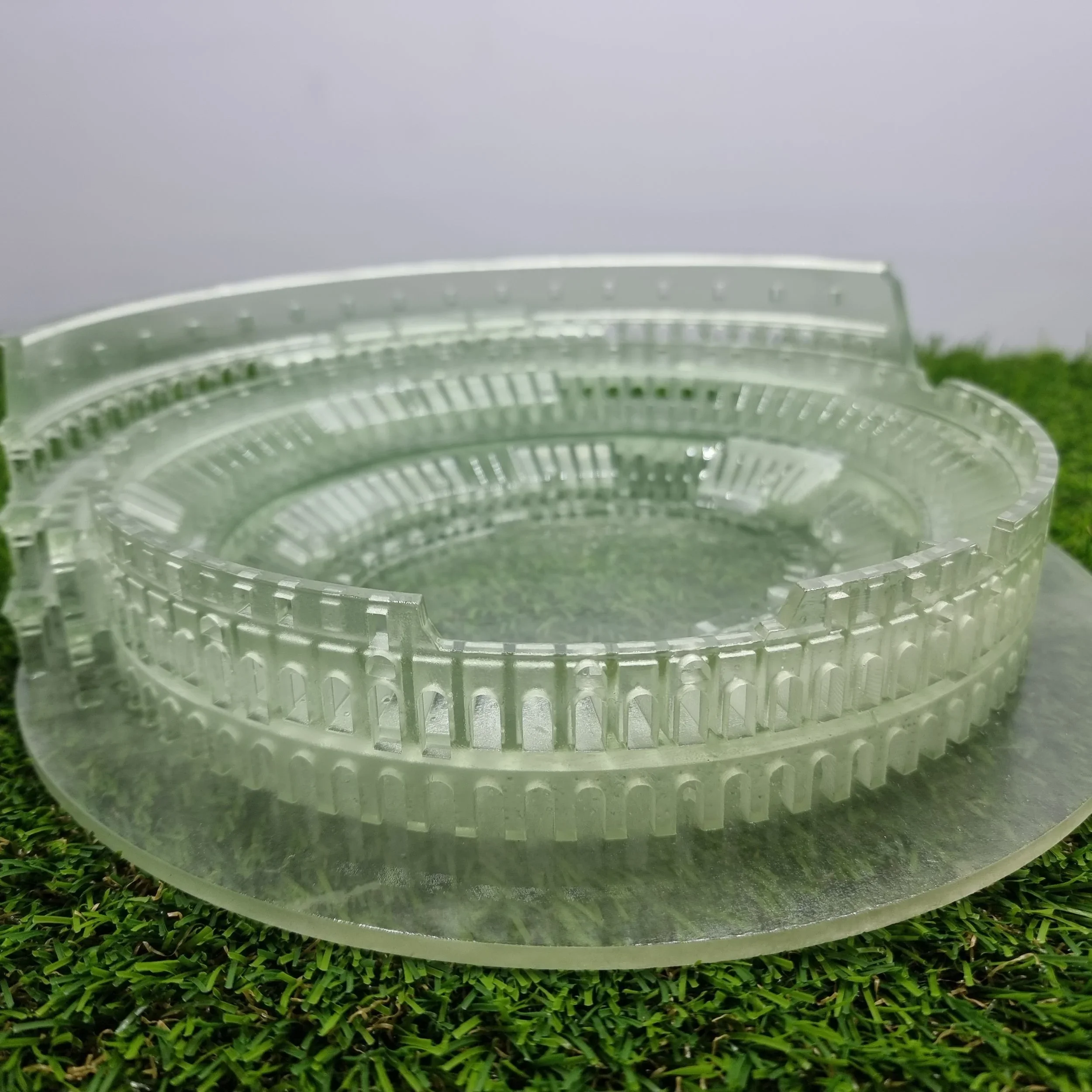 Close-up of clear plastic circular object with multiple small rectangular openings, placed on artificial green grass with a foggy gray sky in the background.