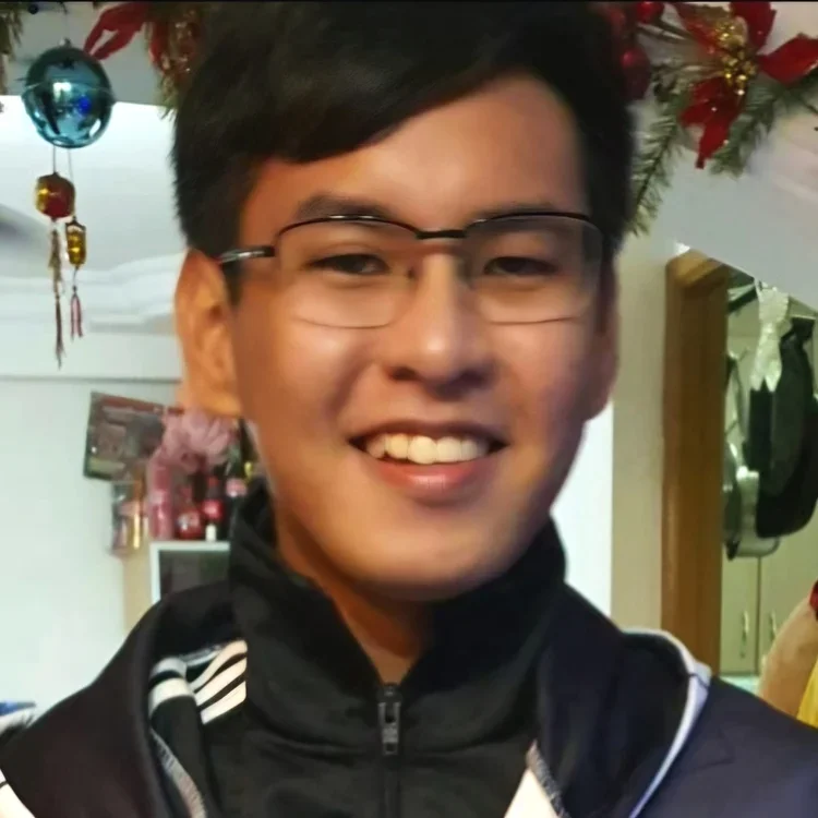 A young man with glasses and short dark hair smiling, wearing a black zip-up jacket, decorated with Christmas ornaments and decorations in the background.