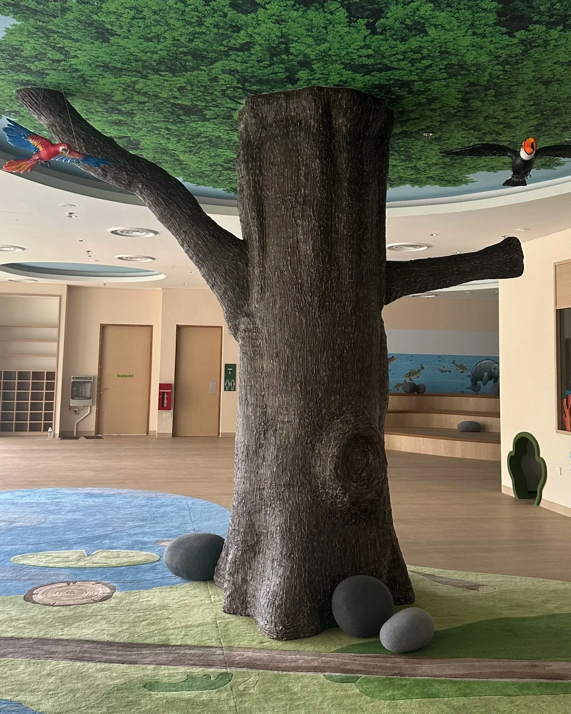 Indoor play area with a large artificial tree and a colorful mural of birds hanging from the ceiling.