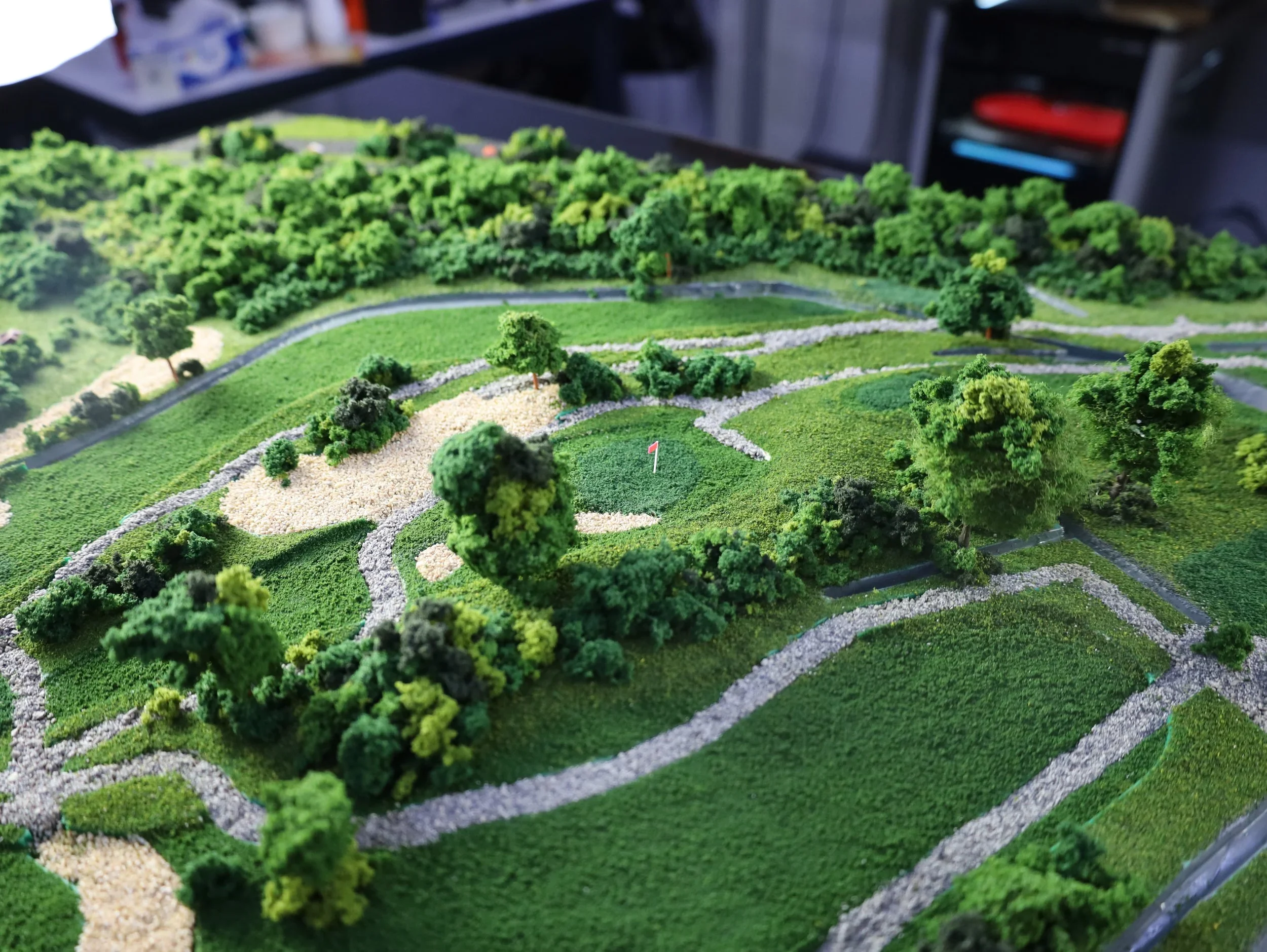 A detailed miniature model of a landscapes with winding paths, green grass, and small trees, resembling a golf course or park.