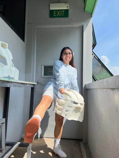 A young woman with glasses and long brown hair, wearing a light blue shirt, light gray shorts, white socks, and white sneakers, is smiling while holding a white plastic chair. She is outdoors on a balcony or terrace with a clear blue sky visible in the background, and an exit sign is above a door behind her.