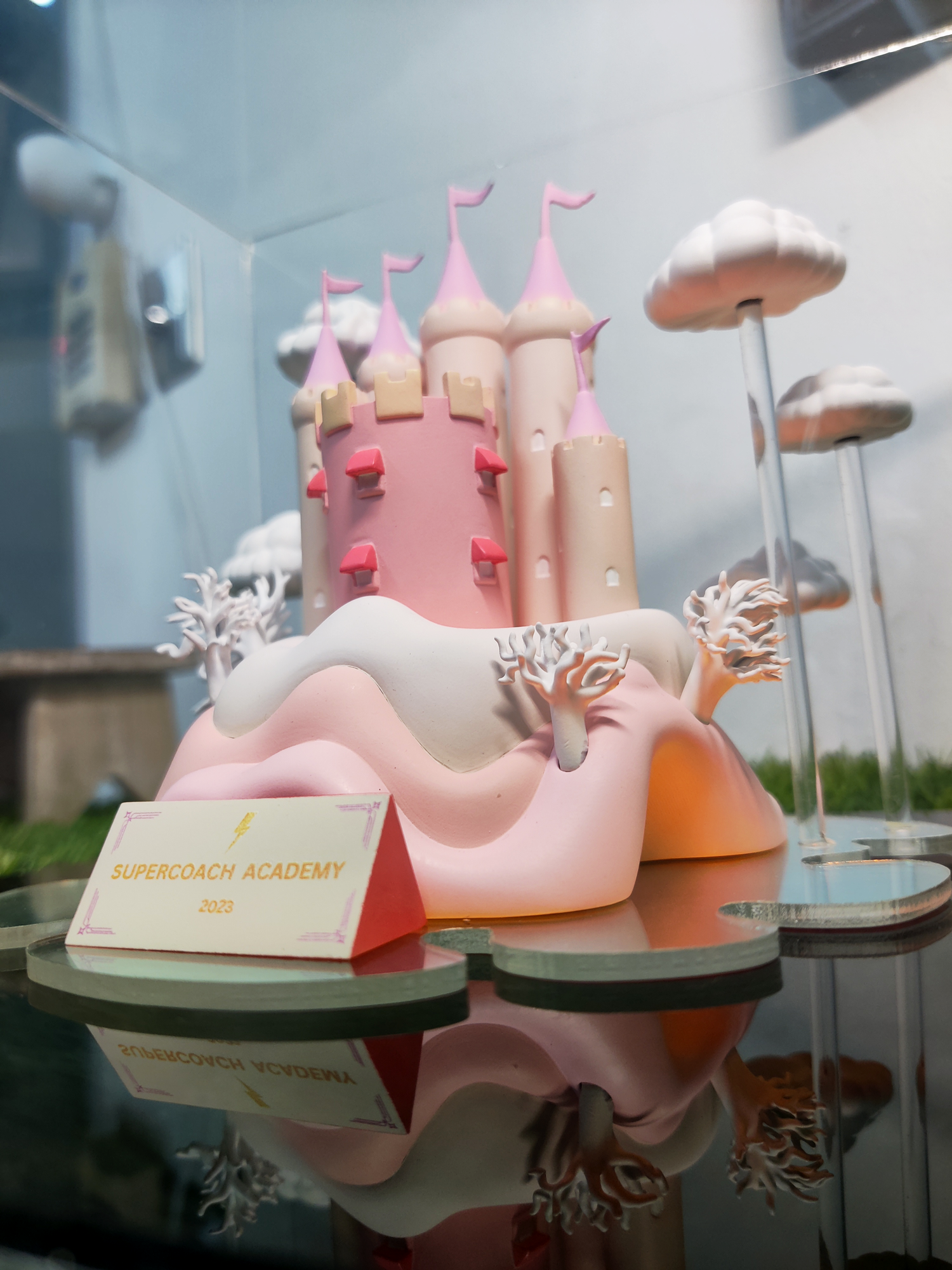 A pink and white castle cake with towers and flags on a glass display case. There are cloudy decorations and a sign that reads 'Supercoach Academy 2023'.