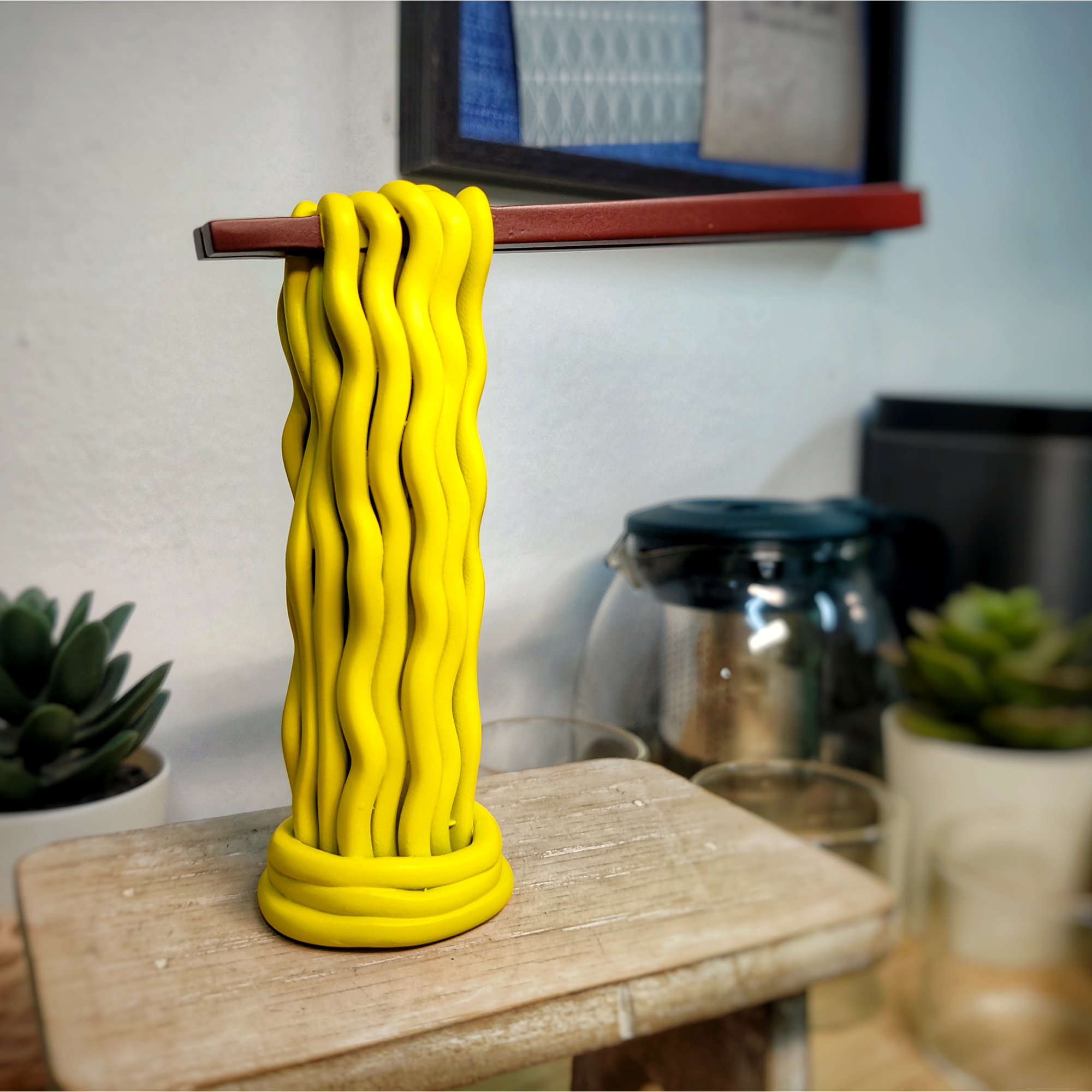 Yellow rubberized pasta hanging on a wooden stick variety of noodles on a wooden surface with plants and kitchen appliances in the background.
