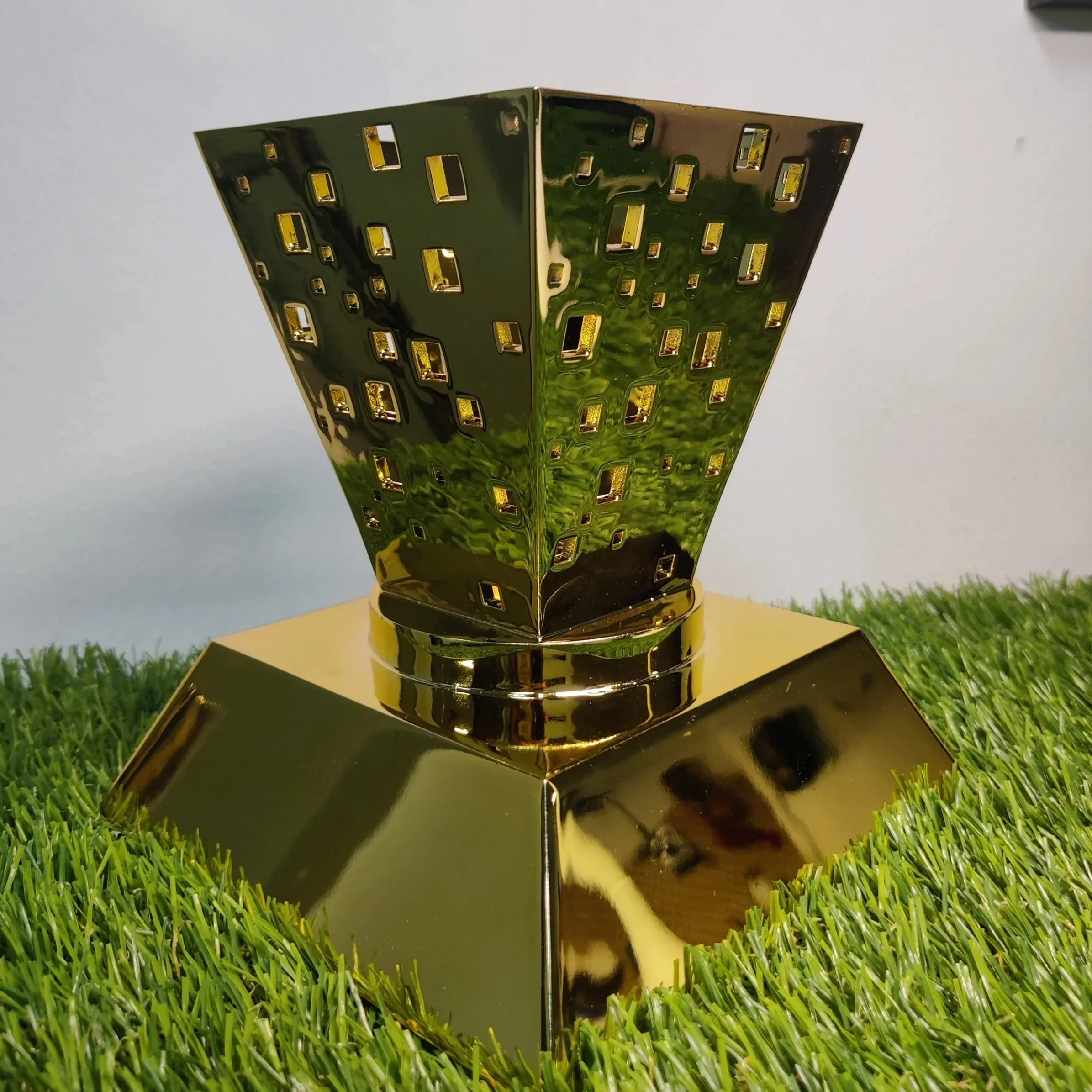 A shiny gold vase with a perforated design sits on a gold base on grass, reflecting the surroundings.
