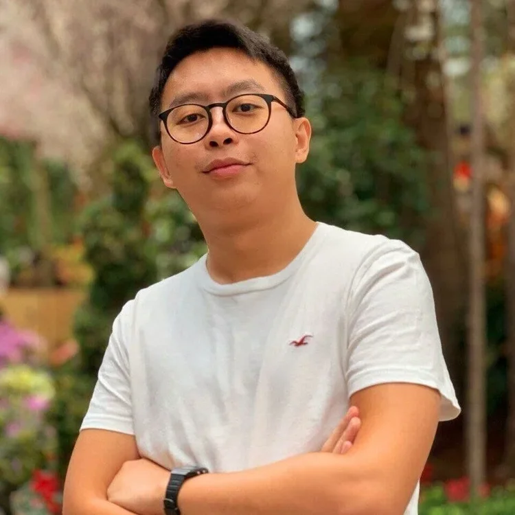 Young man with glasses in a white t-shirt standing outdoors with arms crossed, greenery and trees in the background.