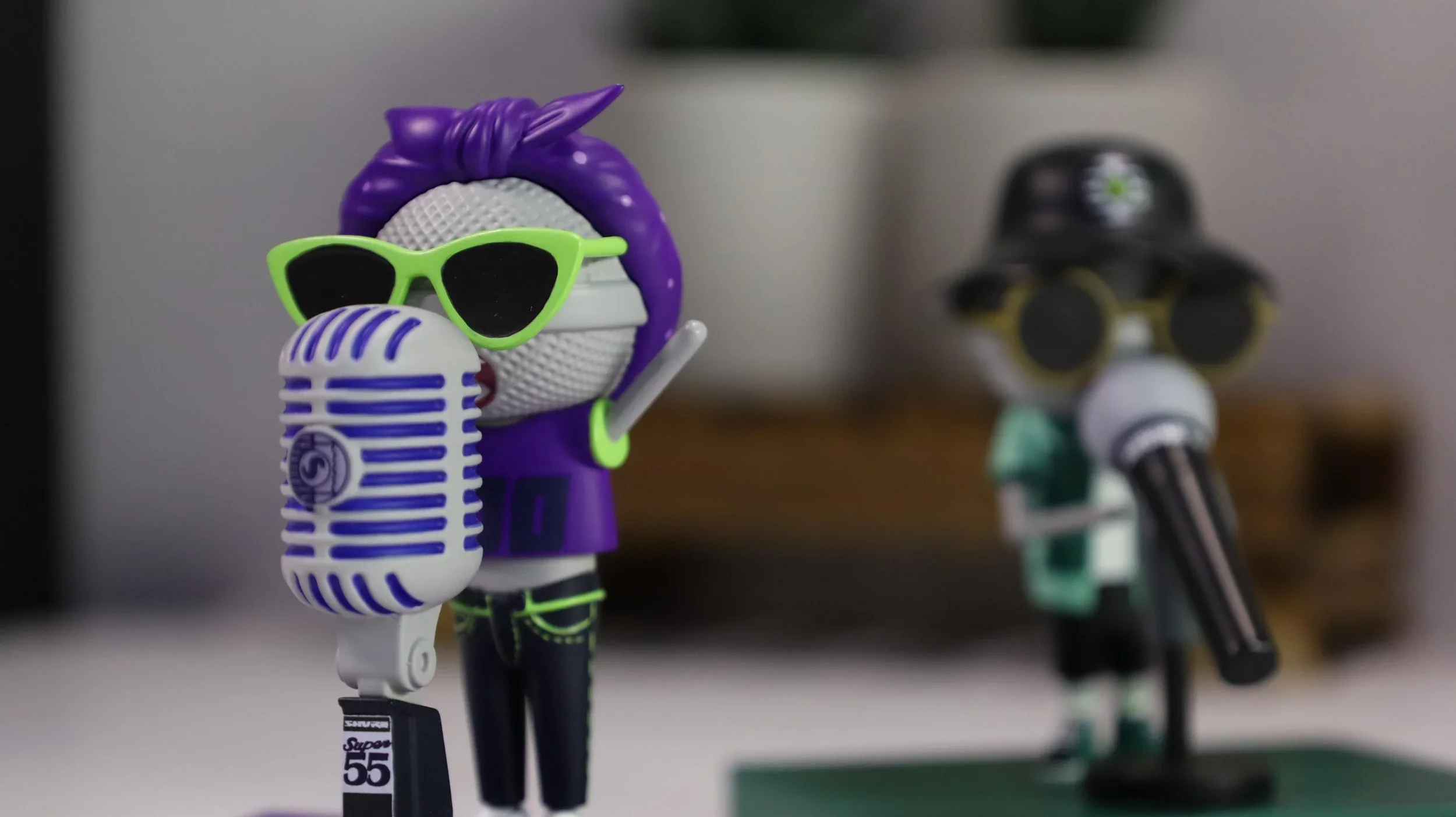 Two cartoon-style figurines with microphones: one with purple hair, green sunglasses, and a purple hoodie, and another with a black hat, green outfit, and large round glasses, both standing on display.