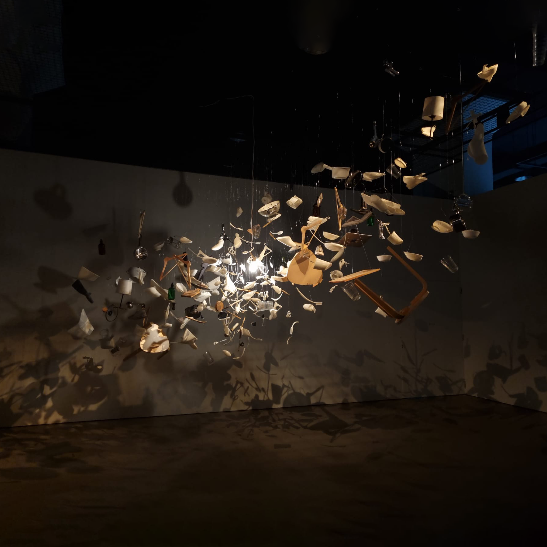 An art installation with various household items like cups, plates, and utensils suspended in mid-air, casting shadows on the wall behind.