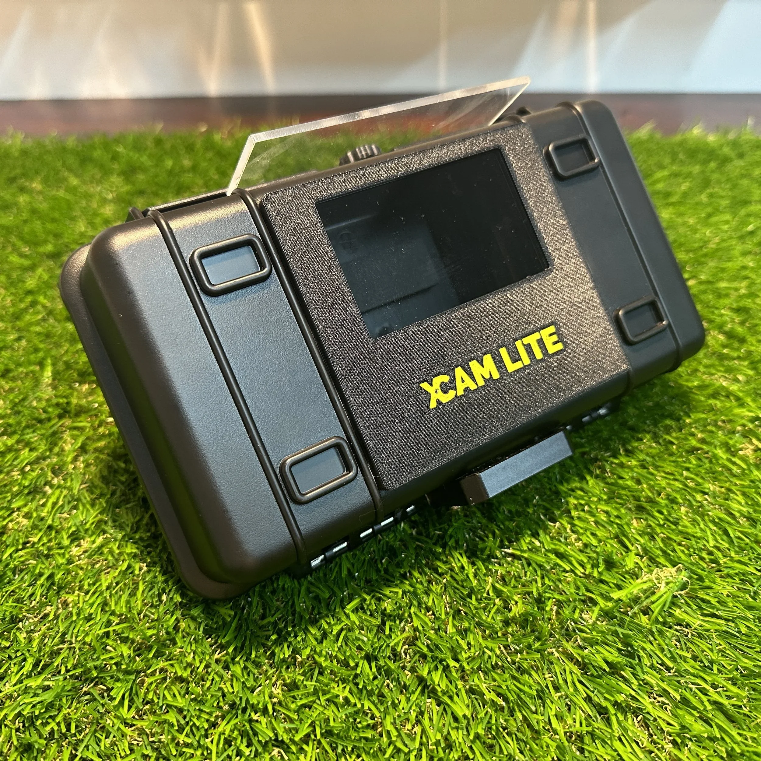 A black device labeled 'XCAM LITE' resting on green artificial grass with a reflective surface in the background.