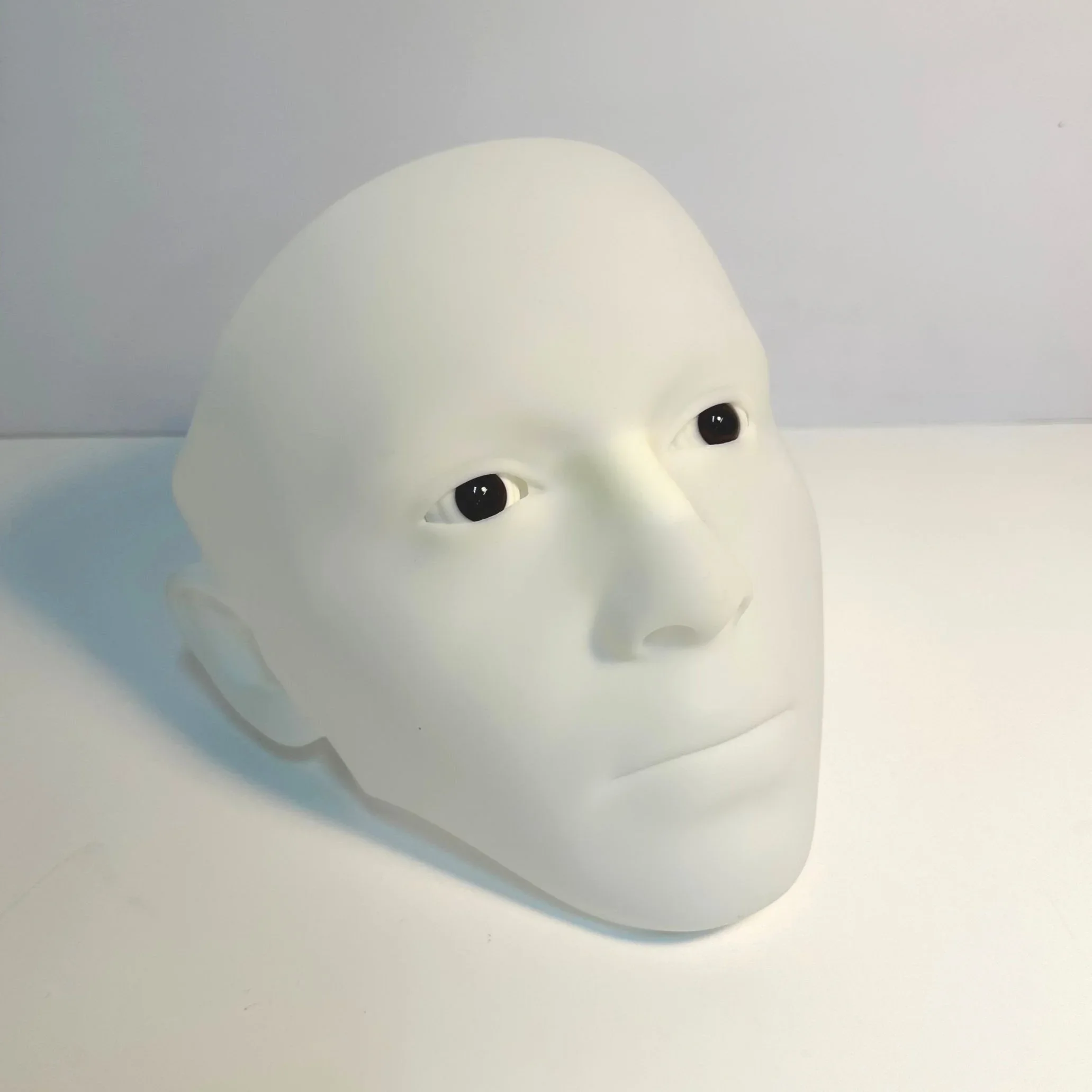 White mannequin head with realistic facial features and black eyes, placed on a white surface against a plain background.