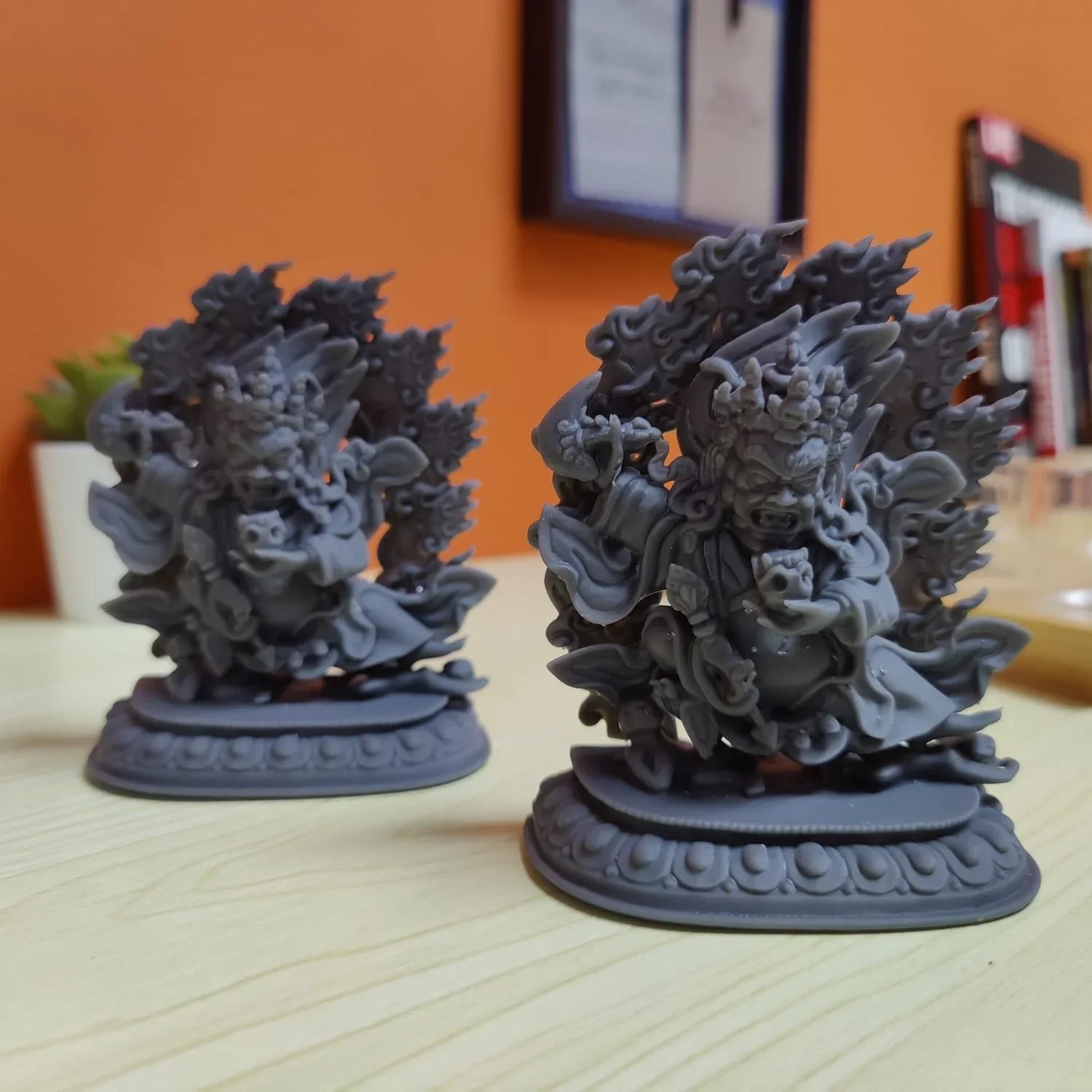 Two detailed gray statues of mythological creatures with fierce faces, surrounded by flames, on oval bases, placed on a light-colored wooden surface.