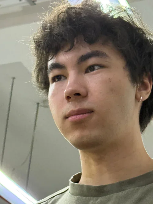 A young man with curly dark hair and fair skin looking thoughtfully to the side in an indoor setting.