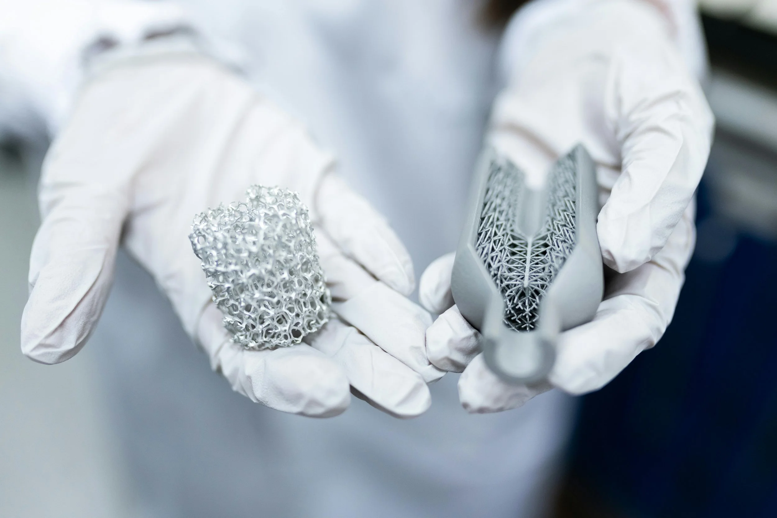 Person wearing white gloves holding metallic 3D-printed lattice objects, including a porous cylindrical piece and a complex geometric structure.