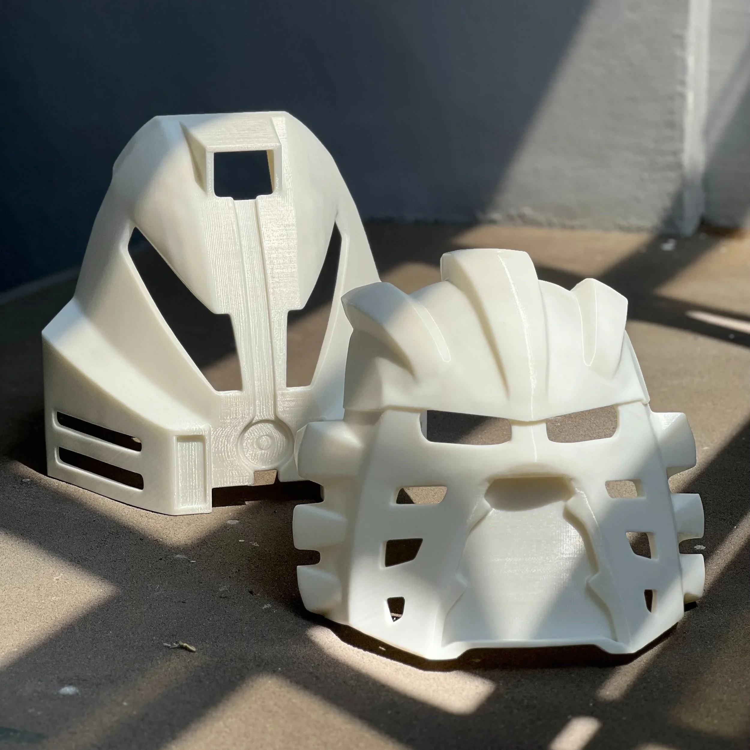 Two white plastic masks resembling robotic or sci-fi characters, one with a more helmet-like design and the other with a more detailed, angular face design, resting on a concrete surface with shadows.