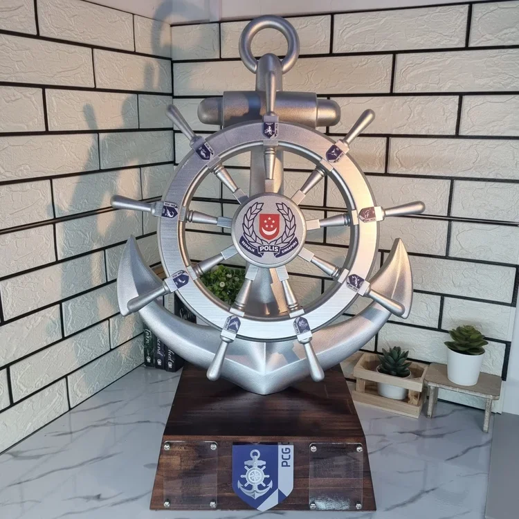 Decorative nautical-themed display featuring a silver ship's wheel mounted on an anchor-shaped base, with police insignia and plaque, placed on a wooden platform on a marble countertop, with potted plants in the background.