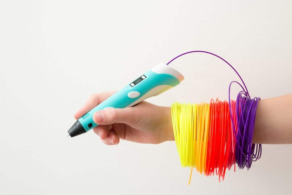 A hand holding a 3D pen, with colorful plastic filament strands wrapped around the wrist in yellow, red, and purple.