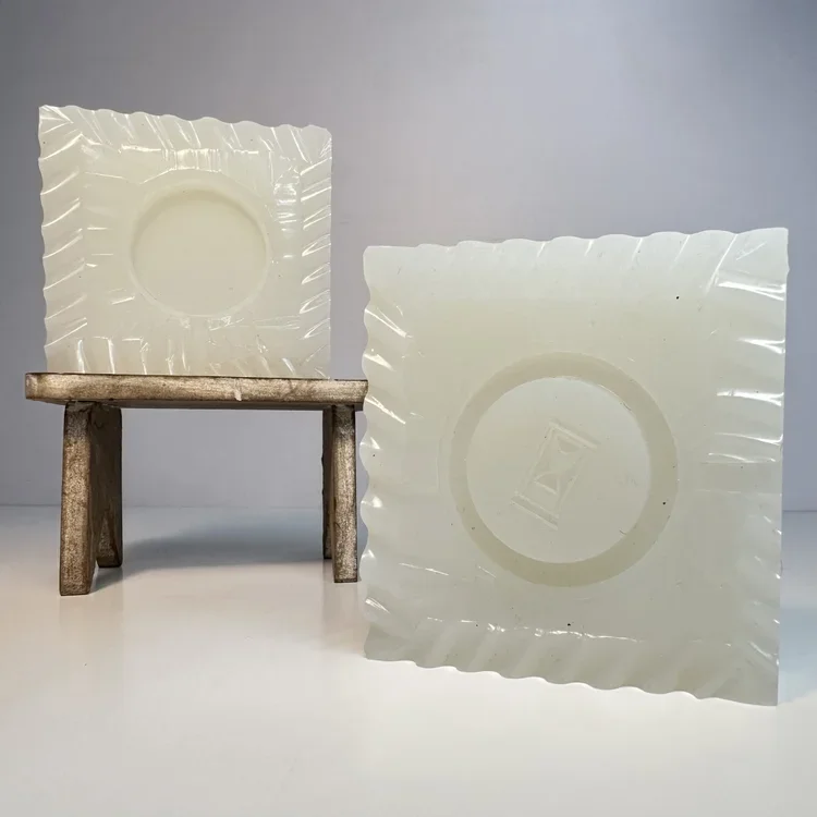Two square white plates with wavy edges and a circular indentation, one placed on a small wooden stool and the other leaning against a light gray wall.