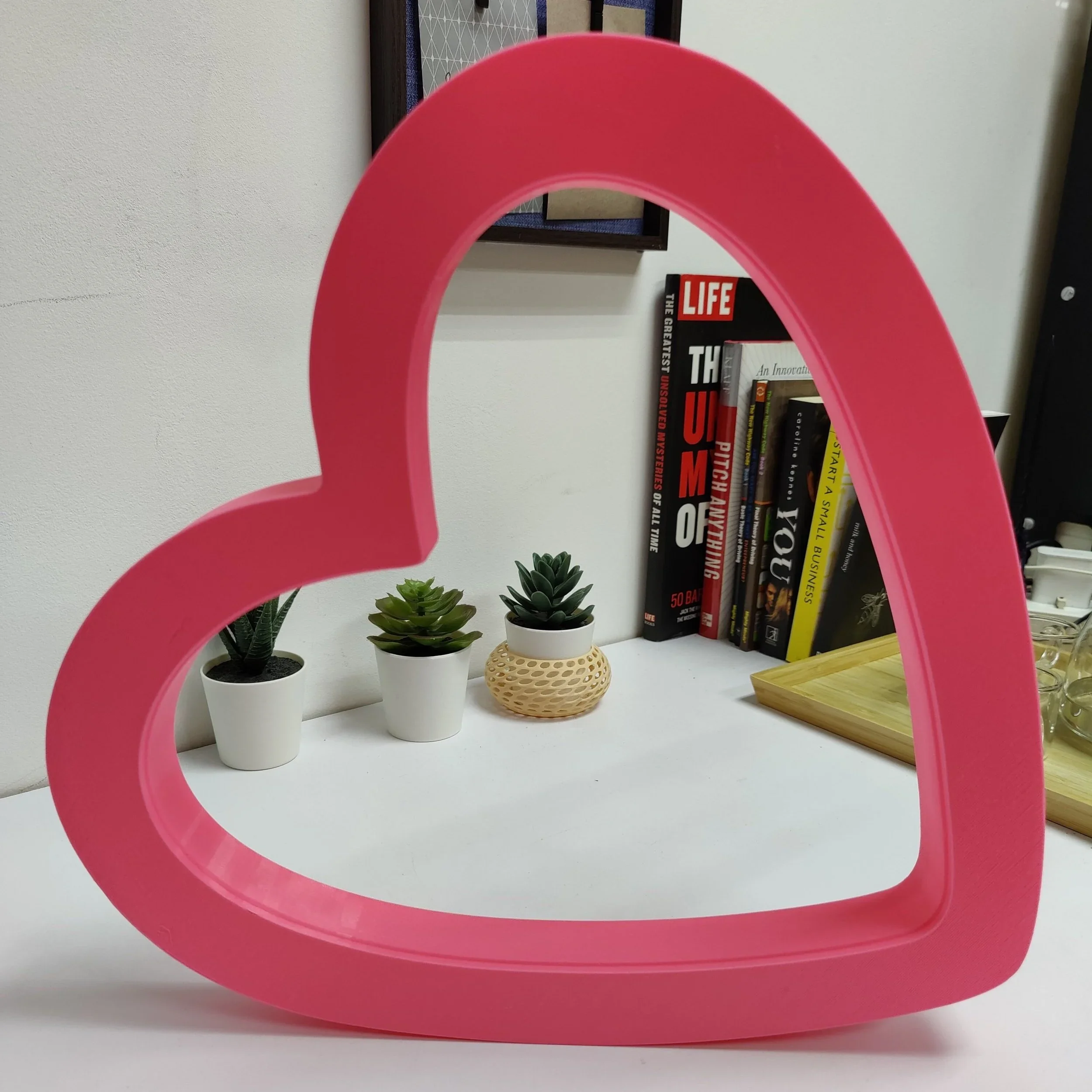 Pink heart-shaped frame in the foreground, with small potted succulent plants, stacked books, and a wooden tray in the background.