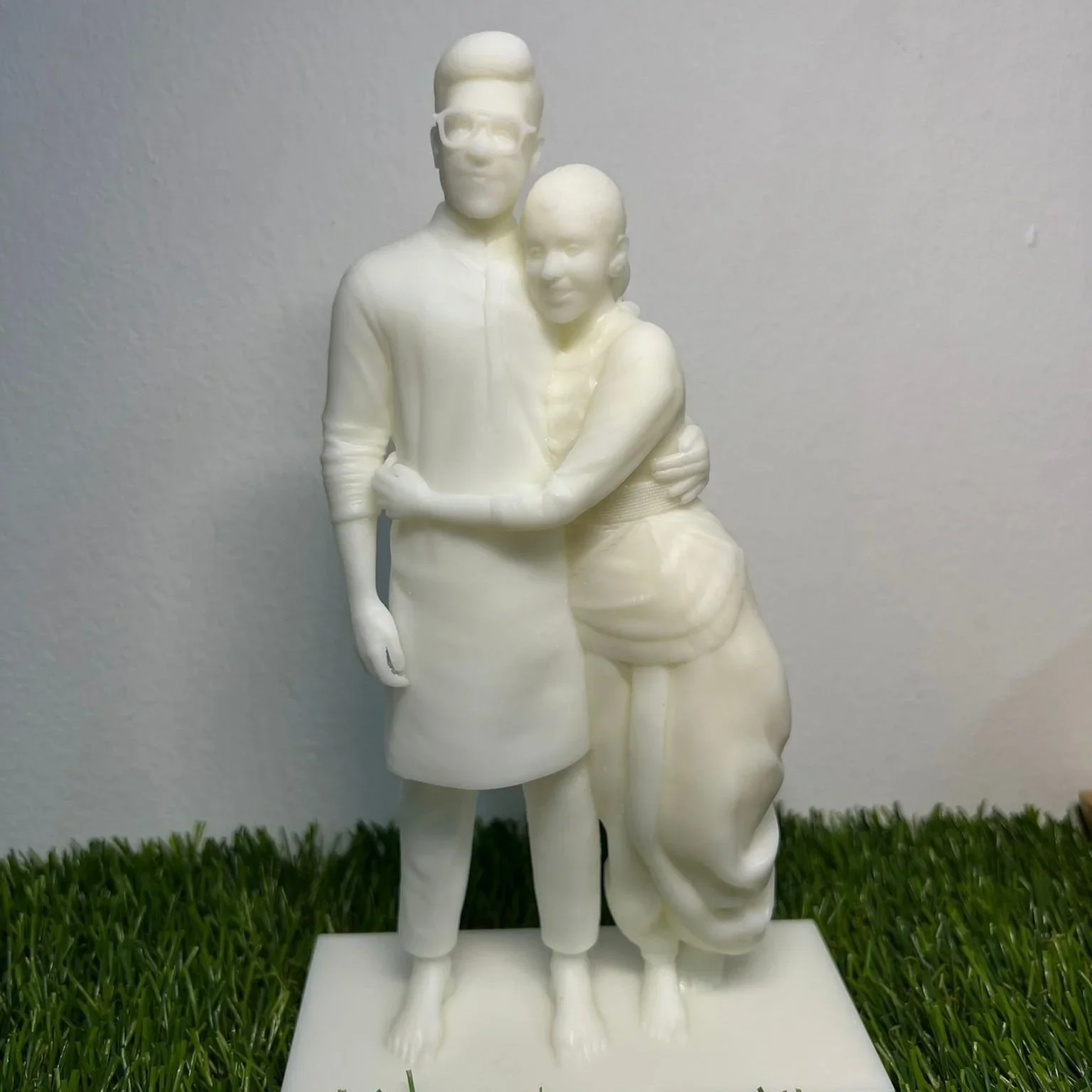 White sculpture of a man and woman standing closely with their arms around each other, on a green grass surface.