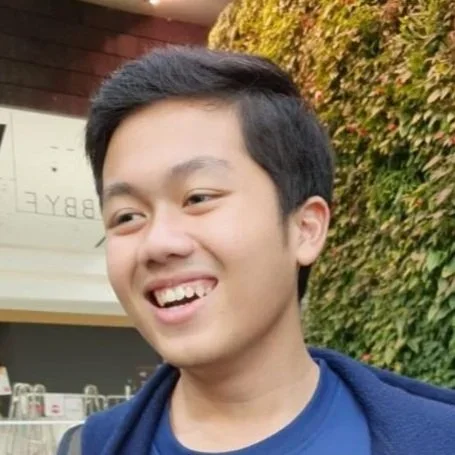 A young man with short dark hair smiling outdoors next to a wall covered with green and red foliage.