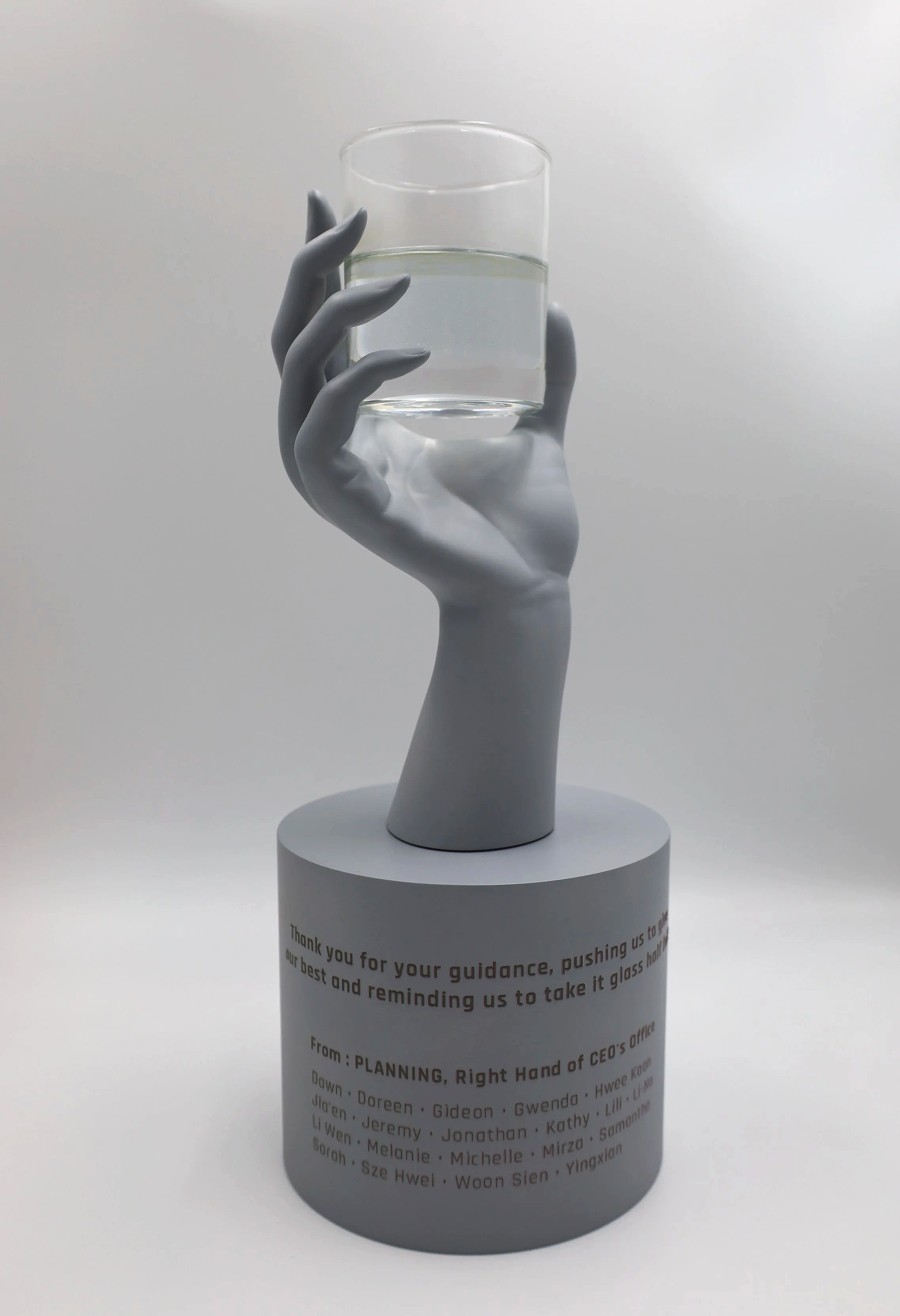 A decorative sculpture of a hand painted in white, holding a glass of water, mounted on a cylindrical base with a message and list of names printed on it.