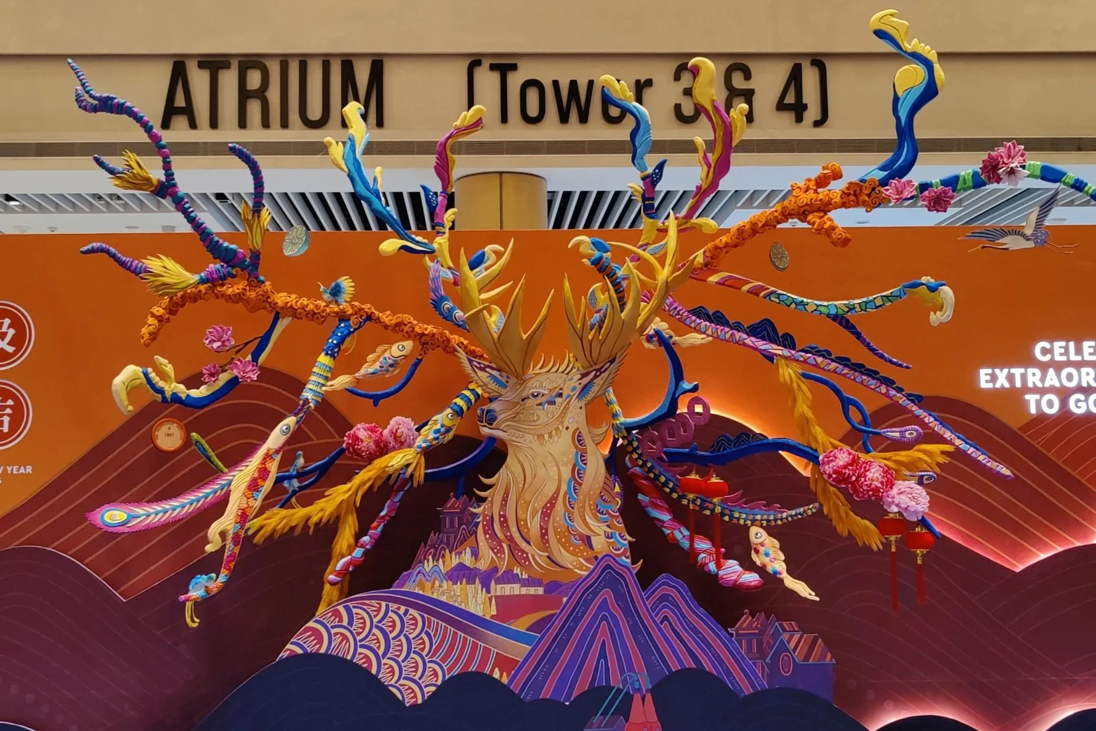 Colorful decorative art installation of a mythical deer with multiple antlers, set against an orange backdrop with text about an atrium and tower numbers.