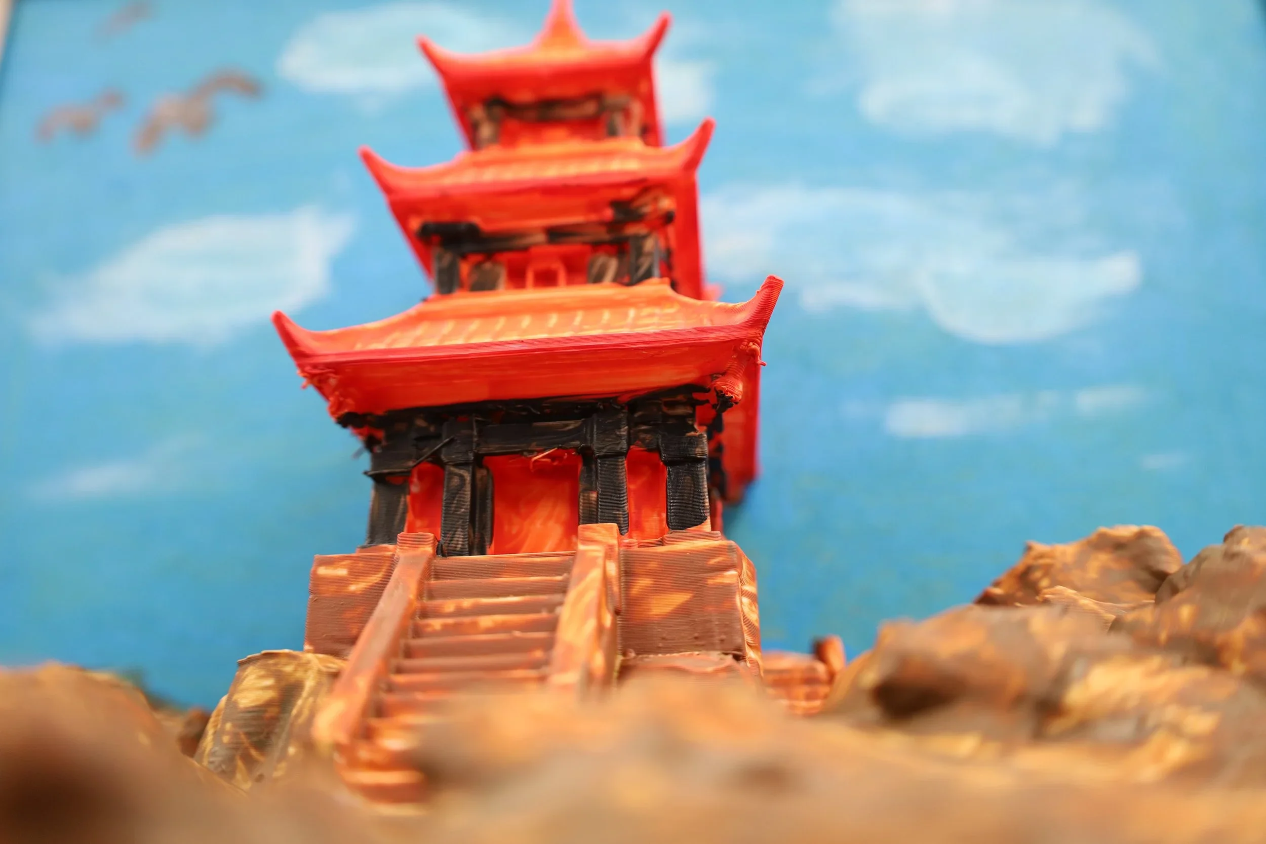 Miniature traditional Chinese pagoda with red and black details, placed on a rocky surface, with a blue sky and clouds in the background.