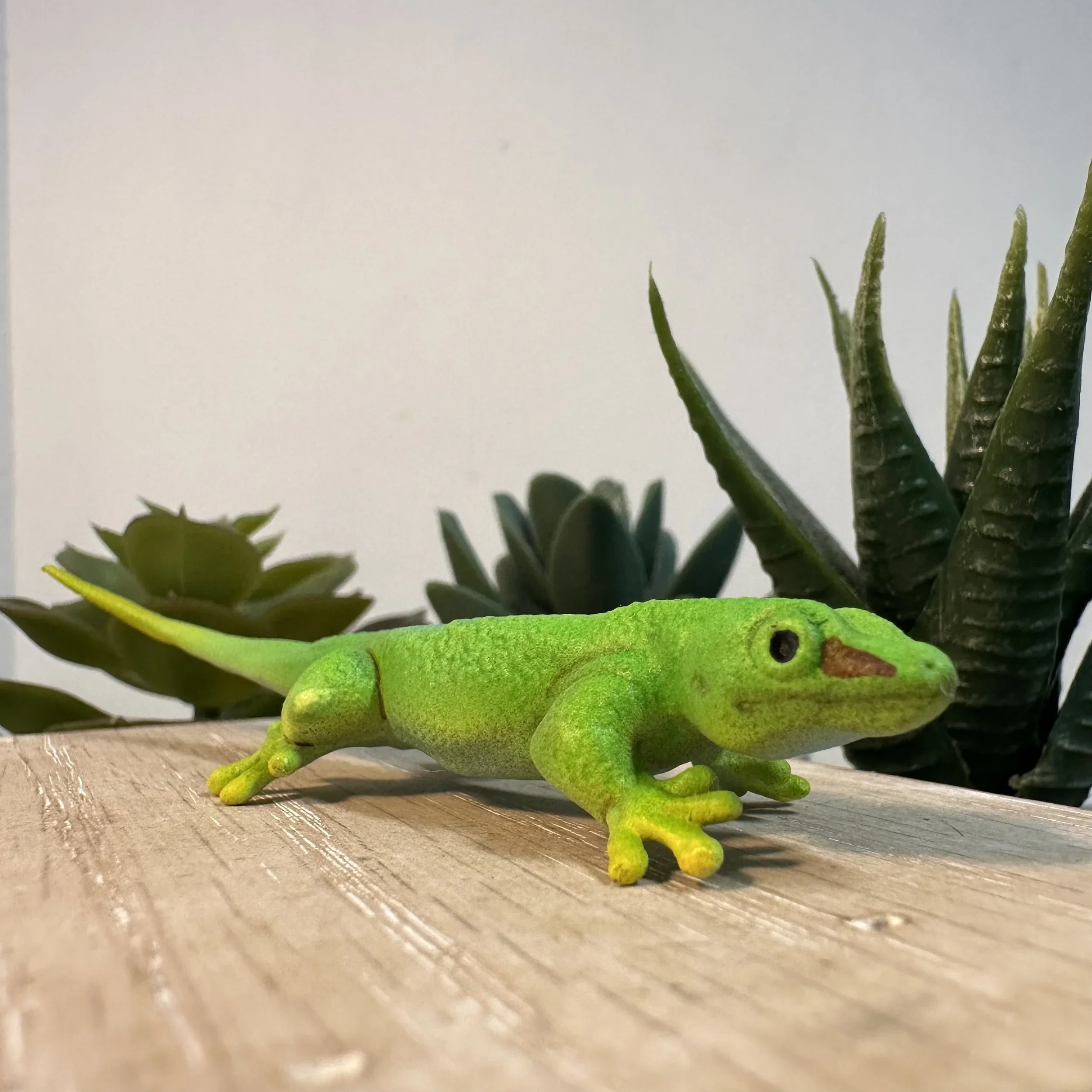 A toy green gecko on a wooden surface with succulent plants in the background.