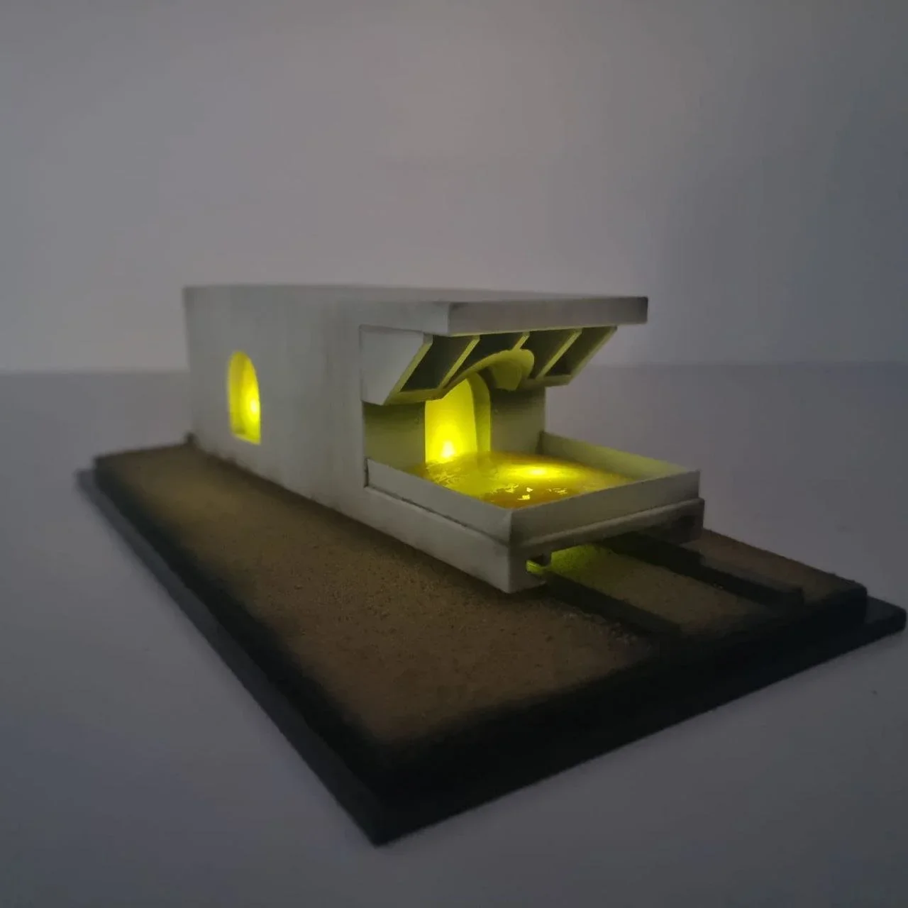 A modern architectural model of a house with illuminated yellow windows and a pond or pool in front, set on a wooden base.