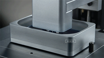 Close-up of a metallic 3D printer printing with a gray filament, positioned on a metallic surface with a black knob nearby.