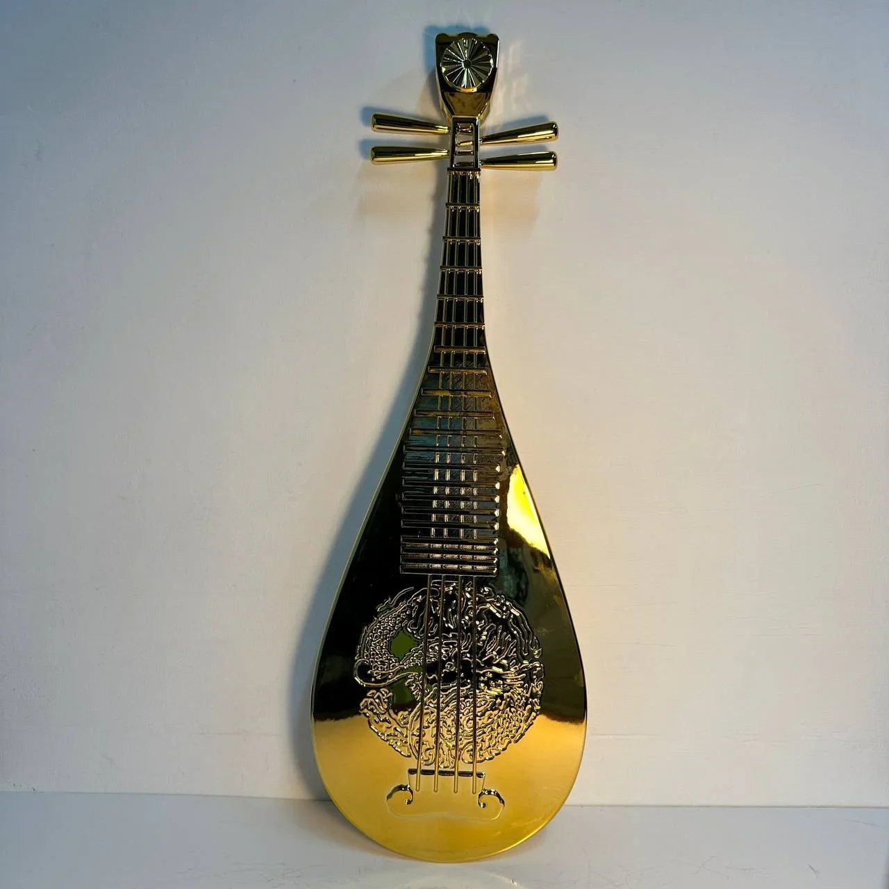A gold-colored, stringed musical instrument shaped like a guitar with decorative engravings, mounted on a white wall.
