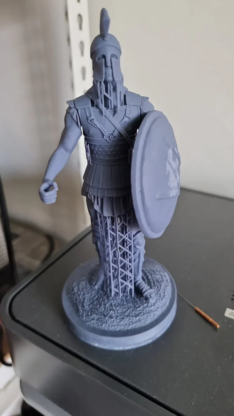 Gray 3D-printed figurine of an ancient Roman or Greek soldier with a helmet, shield, and armor, standing on a circular base.