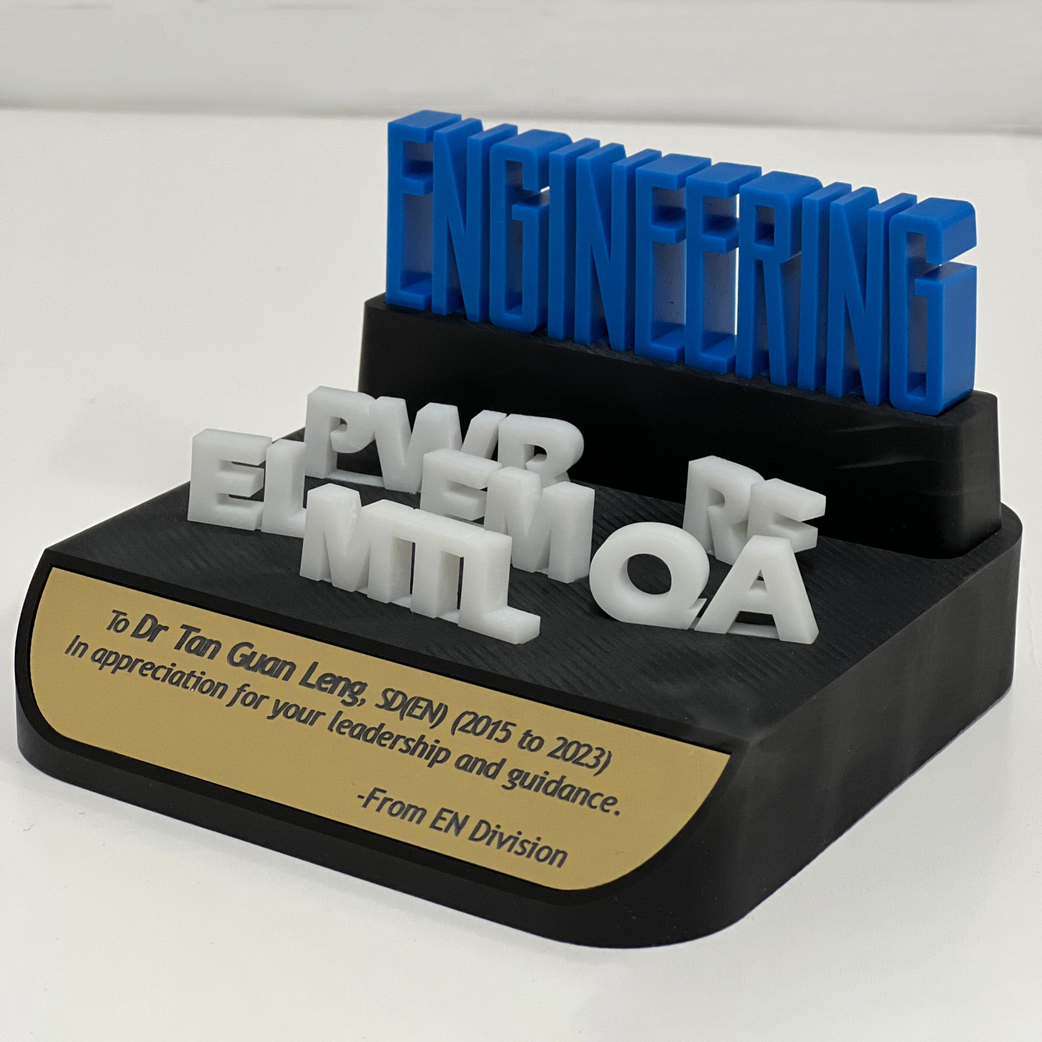 A black display stand with a yellow plaque featuring an appreciation message. Multiple 3D plastic letters spelling 'ENGINEERING', 'PWR', 'ELEC', 'MECH', 'A' are arranged on the stand.