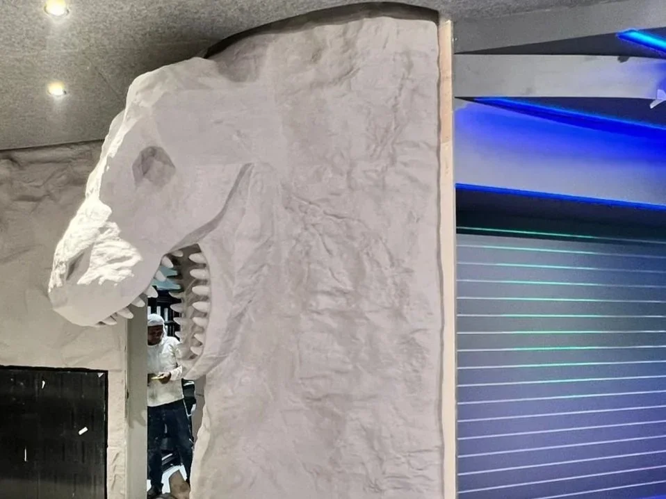 A large, white sculpture of a dinosaur skull, likely a T-Rex, mounted on a wall in an indoor setting.