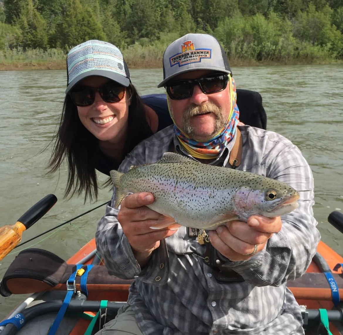 Two people in a boat on a river, one holding a large fish, both wearing sunglasses and hats, surrounded by trees in the background.