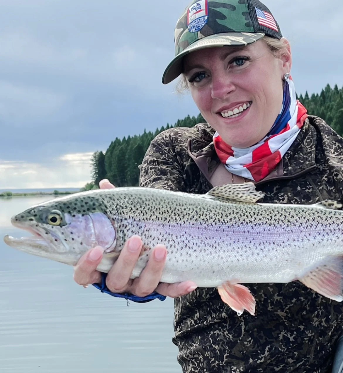 Person wearing a camo hat and jacket, smiling, holding a large fish, likely a trout, by a lake with trees in the background.