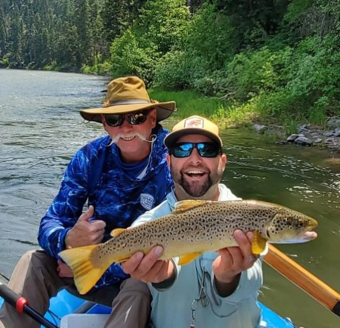 Two men in a boat holding a brown trout near a river with trees in the background.